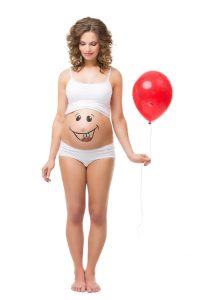 Beautiful girl holding air balloon and looking at her pregnant belly with smily funny face drawn on it. Isolated.